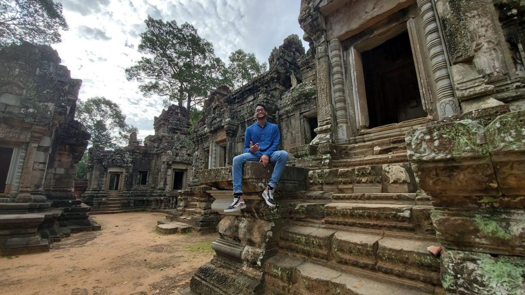 A man sitting on the ancient stone steps of a temple ruin, surrounded by moss-covered walls and weathered architecture.